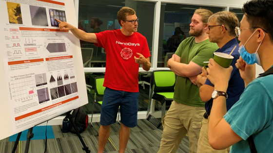 man in red shirt points to poster on easel while others listen to his presentation